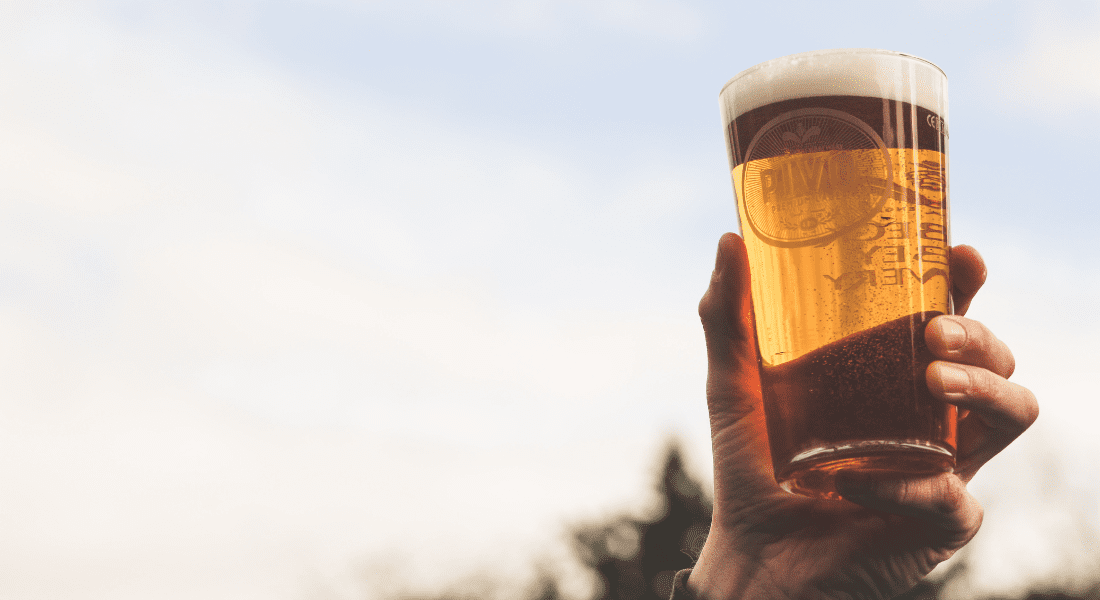 A hand holds up a pint of beer against a cloudy sky background.